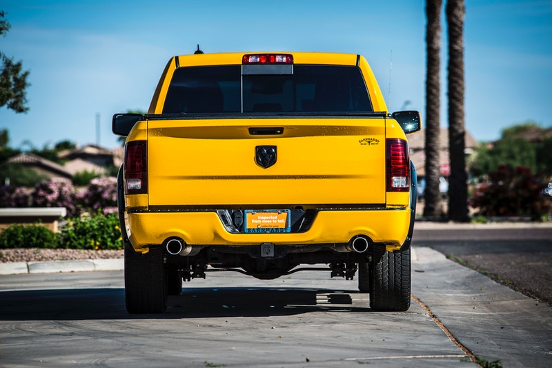 A bright yellow Ram 1500 pickup truck with straight pipe exhaust is shown from the rear, parked on a paved street with palm trees and a blue sky in the background.
