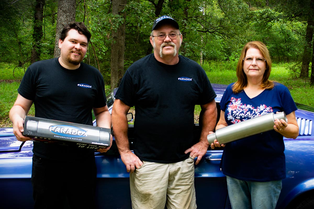 Three owners of Paradox Performance Muffler standing outdoors in front of a blue Ford Mustang, Holding Paradox Performance Muffler products. The center person wears a black shirt with a visible PARADOX Muffler logo, and the parts themselves also show the PARADOX Muffler branding.