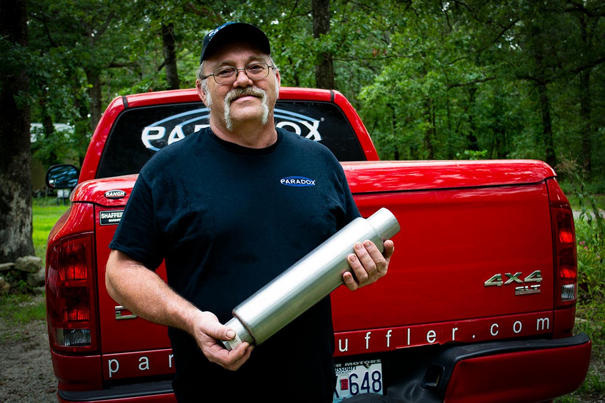 One of the owners of Paradox Performance Muffler holds a muffler in front of his Dodge Ram 1500 equipped with a Paradox Performance Muffler