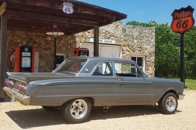 A classic gray Mercury with Paradox Performance Mufflers parked in front of a rustic old Phillips 66 gas station.