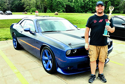 A man stands next to a dark blue Dodge Challenger muscle car with Paradox Performance Mufflers in a paved outdoor lot, holding a trophy and wearing a Paradox Muffler hat.