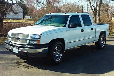 A white Chevrolet Silverado 1500 5.3 pickup truck with a loud muffler by Paradox. It is parked outdoors on a paved surface with trees and a house visible in the background.