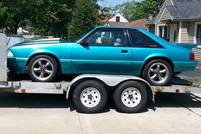 A turquoise blue Fox Body Mustang with Paradox Performance Mufflers on a dual-axle trailer parked outdoors in a driveway with houses and trees in the background.