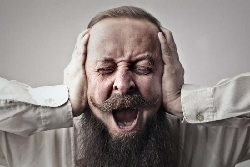 Hearing annoying exhaust drone, a man with a mustache and beard holds both hands to his ears in a gesture of frustration